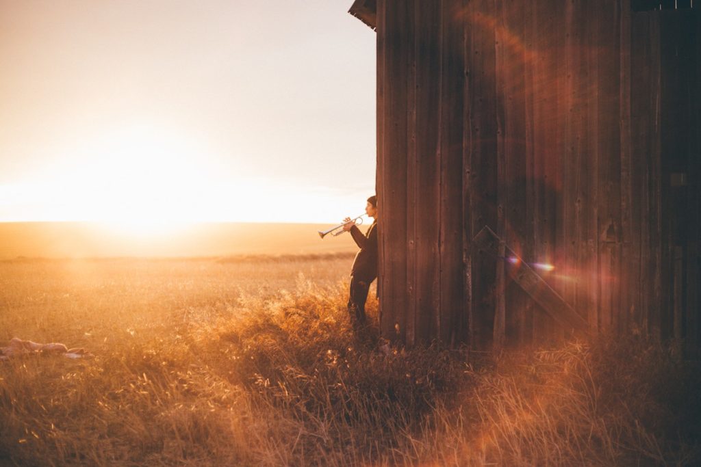 man leaning against sunlit barn playing trumpet