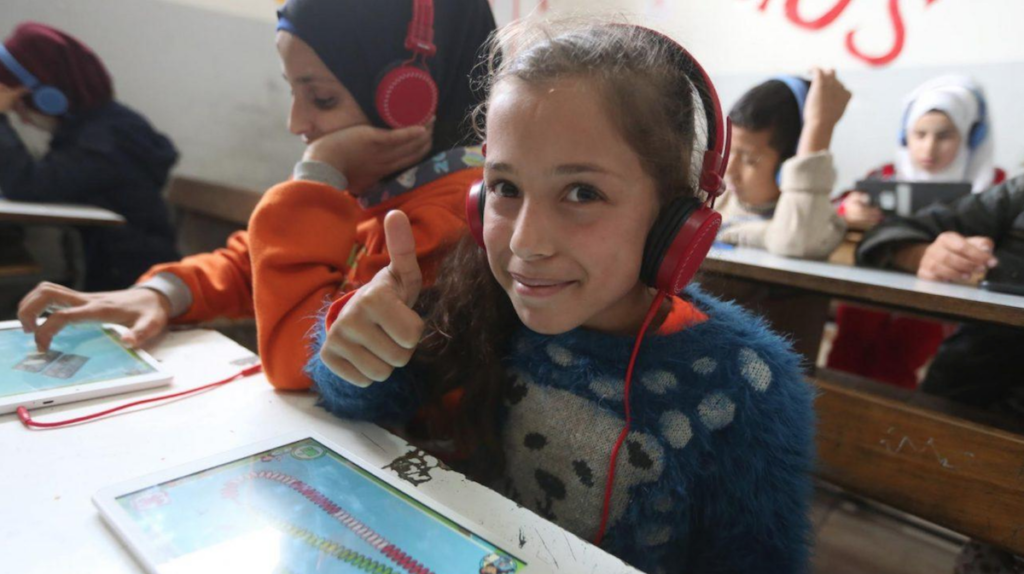 Young girl with headphones on sits at table with tablet computer showing game on screen. Girl smiles and gives a thumbs up to photographer. Other refugee kids in background doing same thing.