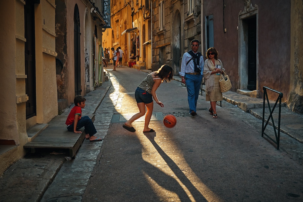 Girl playing with ball in an old part of a city. Boy sits watching and couple walks by.