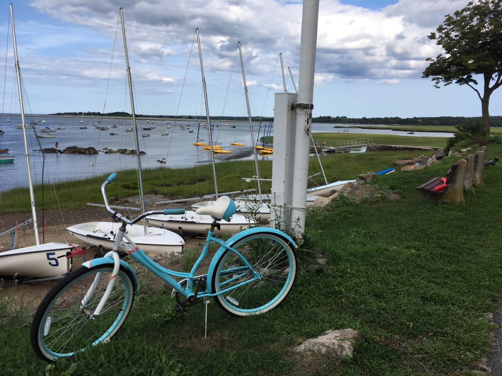 photo looking out to Conomo Point Harbor with many boats moored, in foreground beached small sailboats, and a blue bicyle